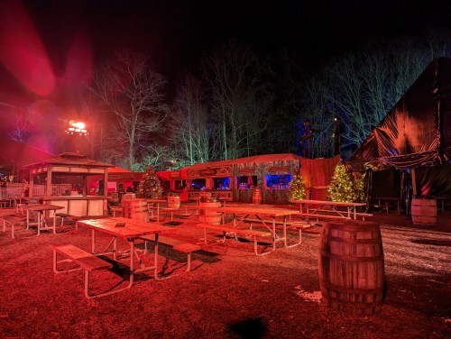 A nighttime scene of an outdoor venue with picnic tables, barrels, and festive decorations under colorful lights.