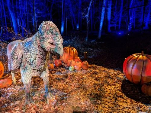 A large, colorful creature resembling a bird stands among pumpkins in a dimly lit forest with blue lighting.