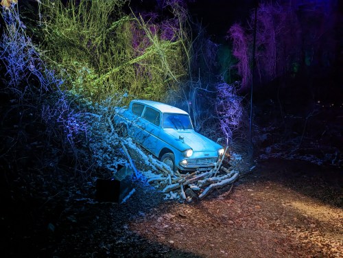 A vintage blue car partially hidden in a forest, surrounded by branches and illuminated by colorful lights.