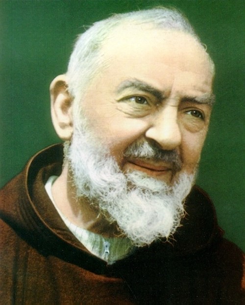 A close-up portrait of a smiling man with a white beard, wearing a brown robe, against a green background.
