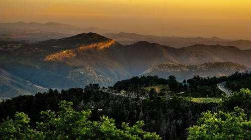 Sunset over mountains with layered peaks, lush greenery in the foreground, and a winding road in the distance.