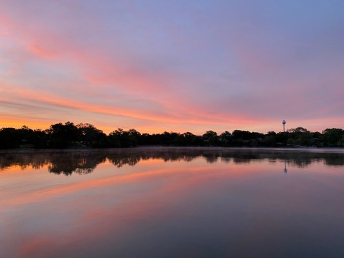A serene lake at sunset, reflecting colorful clouds and trees, with a water tower visible in the distance.