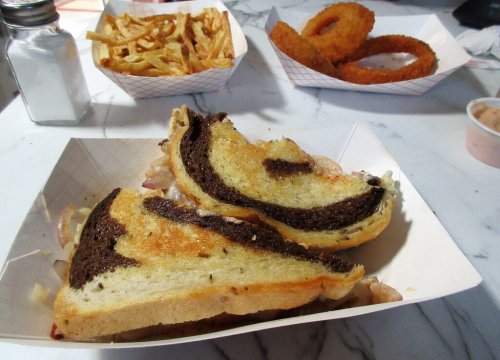 A plate with a grilled sandwich, golden fries, and onion rings in the background on a marble table.