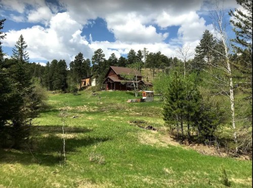 A scenic view of a grassy landscape with two wooden houses surrounded by trees and a blue sky with clouds.