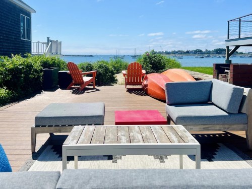 Outdoor seating area with modern furniture overlooking a calm waterfront and boats in the distance. Bright, sunny day.