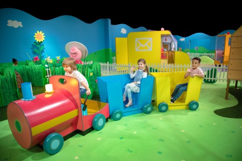 Children playing on a colorful toy train in a vibrant, themed play area with flowers and cartoon scenery.