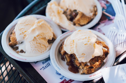 Three bowls of warm apple pie topped with vanilla ice cream, served on a tray with plastic forks.