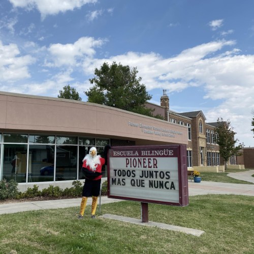 A school building with a sign reading "Pioneer" and a person in a costume standing in front, under a blue sky.