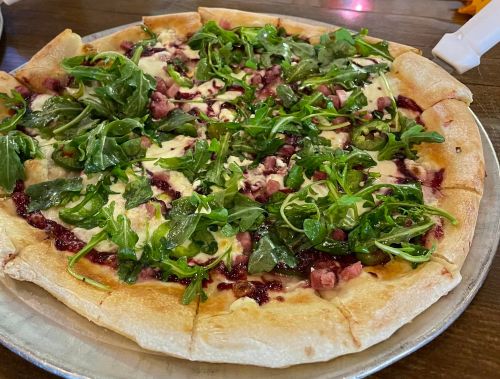 A pizza topped with arugula, ham, and a drizzle of sauce on a golden crust, served on a metal tray.