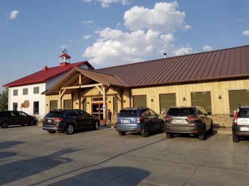 A rustic building with a wooden facade and a red roof, surrounded by parked cars under a blue sky with clouds.