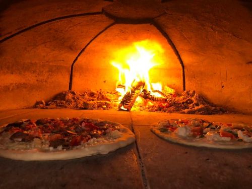 Two pizzas baking in a wood-fired oven with a bright flame in the background.