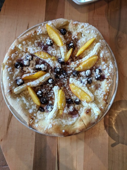 A dessert pizza topped with orange slices, blueberries, and powdered sugar on a wooden table.