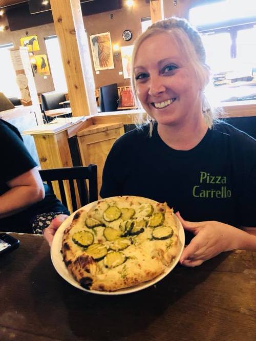 A smiling woman holds a pizza topped with pickles in a cozy restaurant setting.