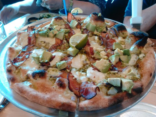 A pizza topped with avocado, lime, pesto, and crispy bacon on a metal serving tray.