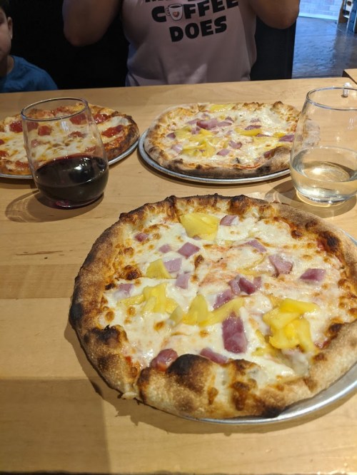 Three pizzas on a wooden table, topped with cheese, ham, and pineapple, alongside glasses of red wine and water.