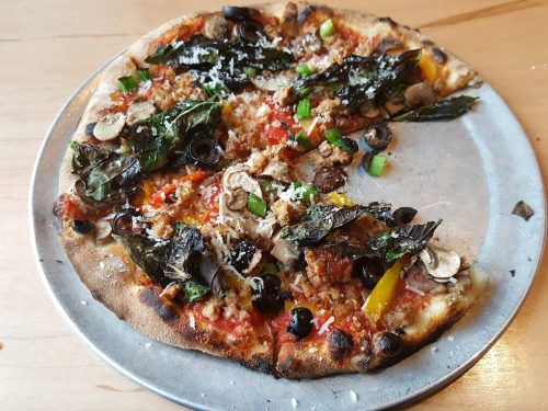 A partially eaten pizza topped with olives, mushrooms, peppers, herbs, and cheese on a metal tray.