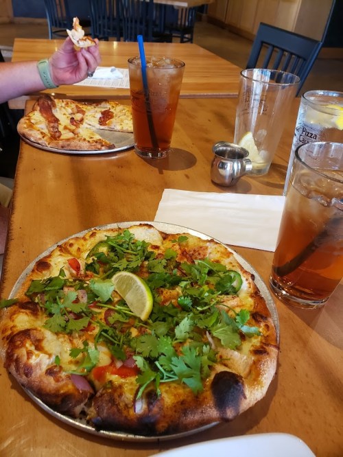 A table with two pizzas, one topped with vegetables and cilantro, and drinks in glasses.