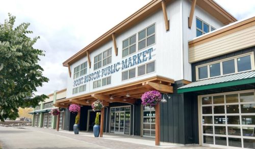 Exterior of Point Ruston Public Market, featuring large windows, wooden beams, and hanging flower baskets.
