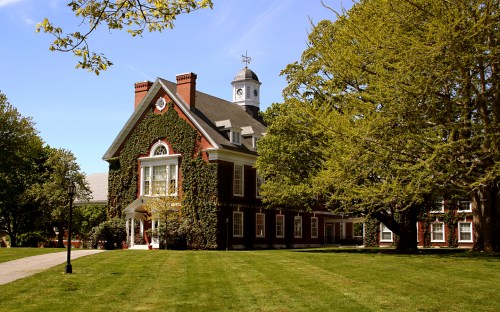 A historic brick building covered in ivy, surrounded by lush green grass and trees under a clear blue sky.