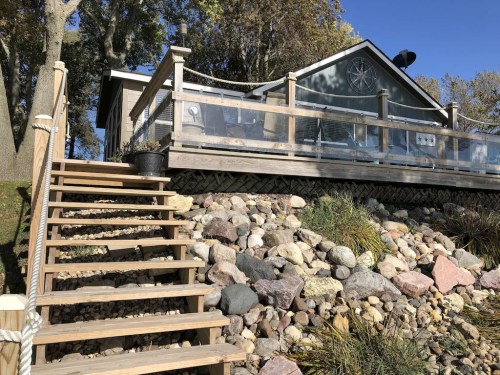 A wooden staircase leads up to a house with a deck, surrounded by rocks and trees under a clear blue sky.