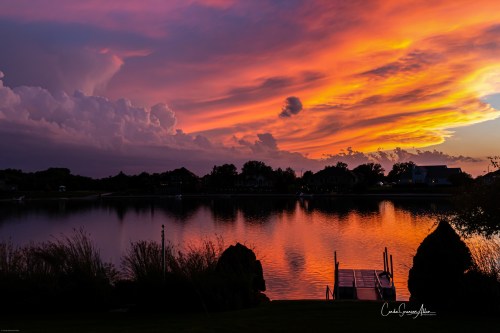 A vibrant sunset over a calm lake, with colorful clouds reflecting on the water and silhouettes of trees and a dock.
