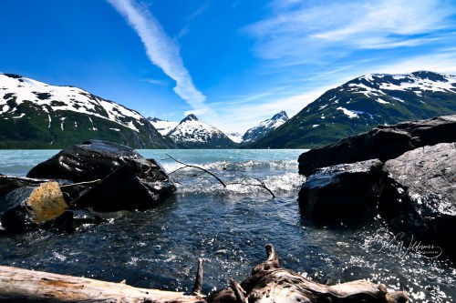 A serene landscape featuring a clear blue sky, snow-capped mountains, and a calm lake with rocky shores.