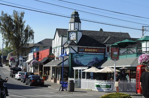 A quaint street scene featuring a bakery, clock tower, and outdoor seating with flowers and shops in a small town.