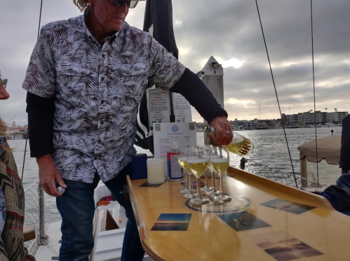 A person pours drinks into glasses on a boat, with a scenic waterfront and cloudy sky in the background.