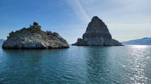 Two rocky formations rise from calm blue waters under a clear sky, with distant mountains visible in the background.