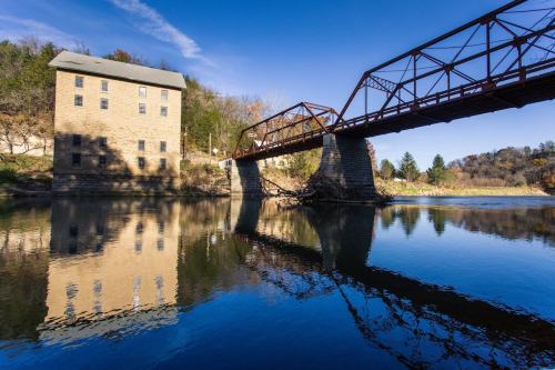 A historic stone building beside a river, with a red metal bridge reflecting in the calm water under a clear blue sky.