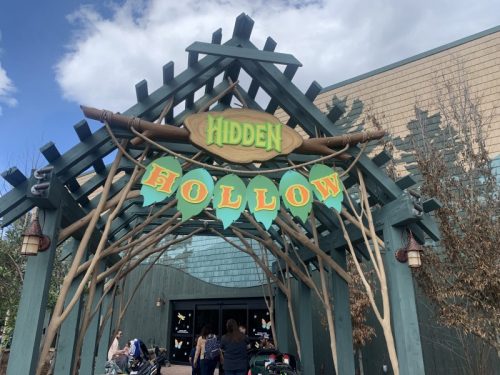 Entrance to "Hidden Hollow" with colorful signage and a wooden archway, surrounded by trees and a blue sky.