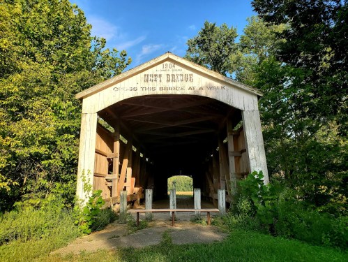 A historic covered bridge surrounded by greenery, with a sign reading "1906" and "Cross this bridge at a walk."