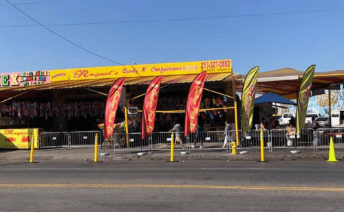 A colorful candy store with flags outside, featuring a bright sign and a fenced area in front.