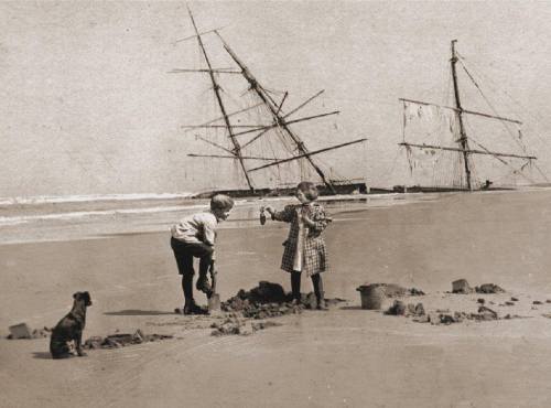 Two children play in the sand on a beach, with a shipwreck and a dog nearby.