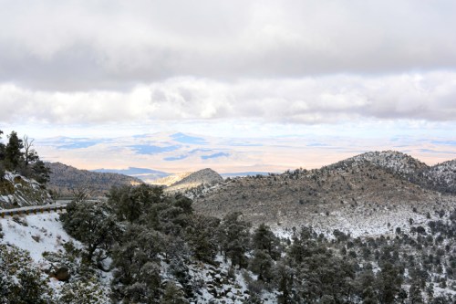 Snow-covered mountains under a cloudy sky, with a distant view of a valley and rolling hills.