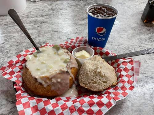 A bowl of clam chowder in a bread bowl, served with a biscuit and a cup of iced soda on a checkered tray.