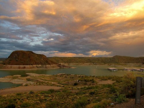 A serene landscape featuring a lake surrounded by hills under a colorful sunset sky with scattered clouds.