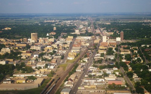 Aerial view of a city with buildings, roads, and greenery stretching into the distance under a clear blue sky.