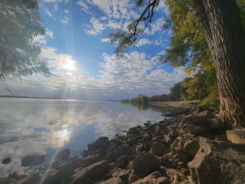 A serene lakeside view with rocky shore, trees, and a bright sky reflecting on the calm water.