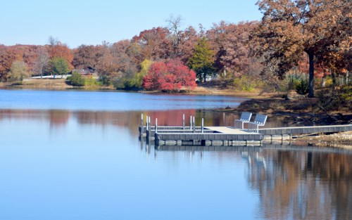 A serene lake scene with a wooden dock, surrounded by autumn foliage and calm water reflecting the clear blue sky.