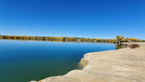 A calm lake reflecting a clear blue sky, surrounded by autumn-colored trees and rocky shoreline.