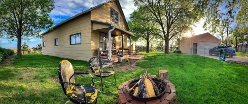 A cozy yellow house with a fire pit and chairs in the foreground, surrounded by green grass and trees.