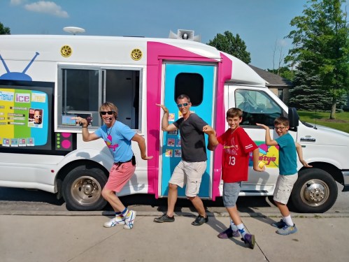 Four people pose playfully in front of a colorful ice cream truck on a sunny day.
