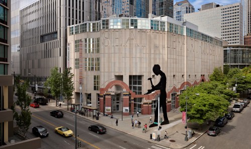 A view of the Seattle Art Museum with a large silhouette sculpture in front, surrounded by city buildings and pedestrians.