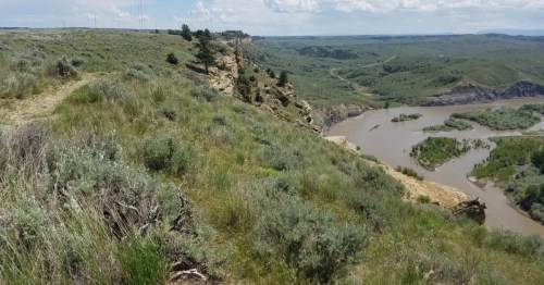 A dirt trail winds its way on top of a mountain ledge toward rocky cliffs. A river and town are visible in the valley below.