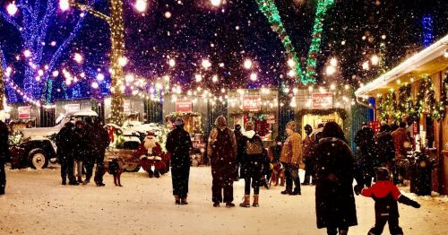 A snowy scene with people enjoying a festive market, decorated with colorful lights and a Santa figure in the background.