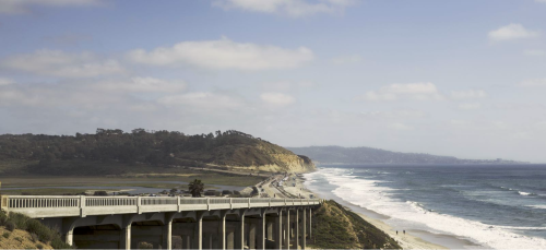 A coastal view featuring a highway along the shore, with waves crashing and hills in the background under a cloudy sky.