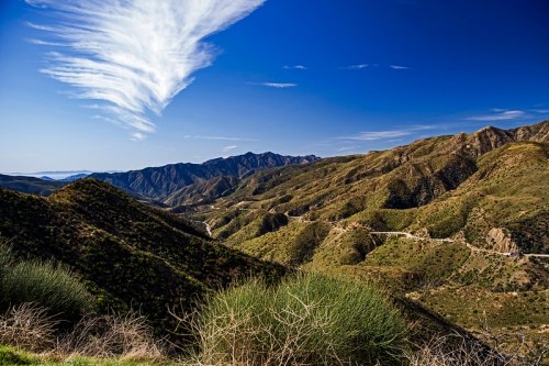 A scenic view of rolling green mountains under a blue sky with wispy clouds, showcasing winding roads in the valley.