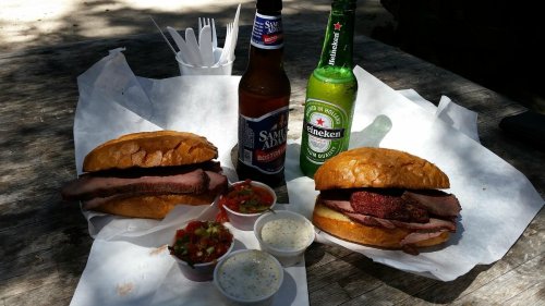 Two sandwiches with meat, two bottles of beer, and dipping sauces on a picnic table.