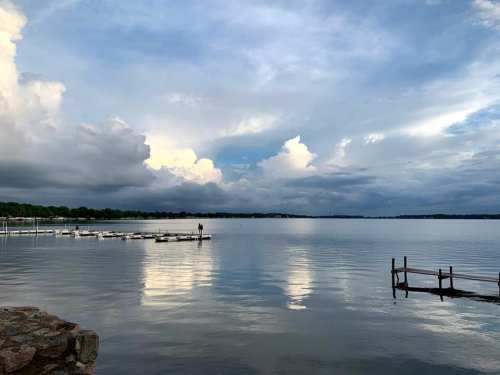A serene lake scene with calm waters reflecting clouds and a distant dock under a partly cloudy sky.